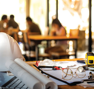 Desk with construction helmet, laptop, glasses and tape measure with students out of focus in the background