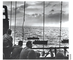 During World War II, U.S. Marines wait aboard a Coast Guard manned combat transport at Tarawa for the invasion barges that will take them ashore. Beyond the rail, Coast Guard coxswains can be seen maneuvering loaded barges.